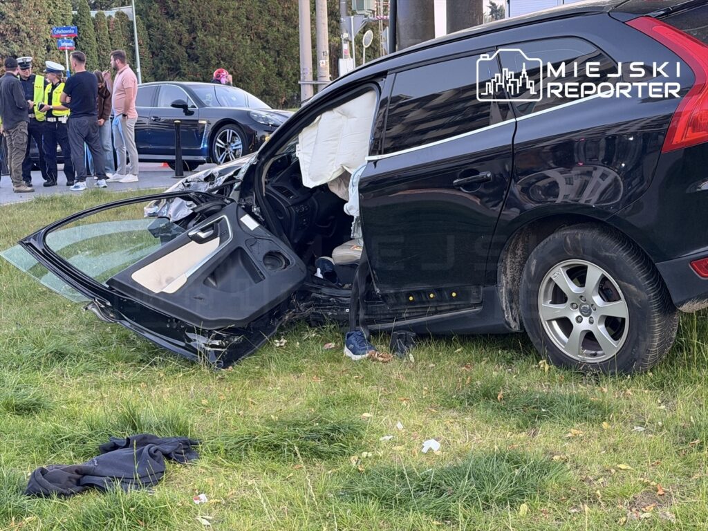 Rozbity czarny samochód z otwartymi drzwiami i wystrzeloną poduszką powietrzną stoi na trawniku, obok policjanci.