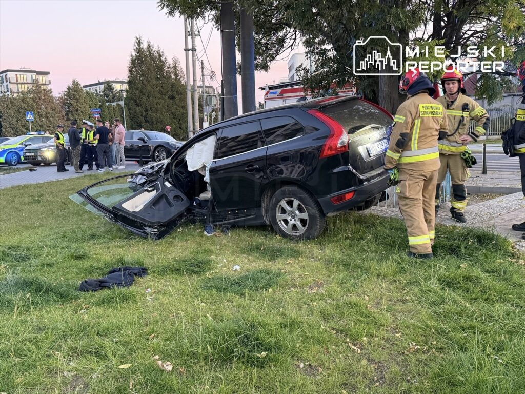 Uszkodzone czarne Volvo stoi na trawniku przy ulicy, obok niego pracują strażacy w odzieży ochronnej.