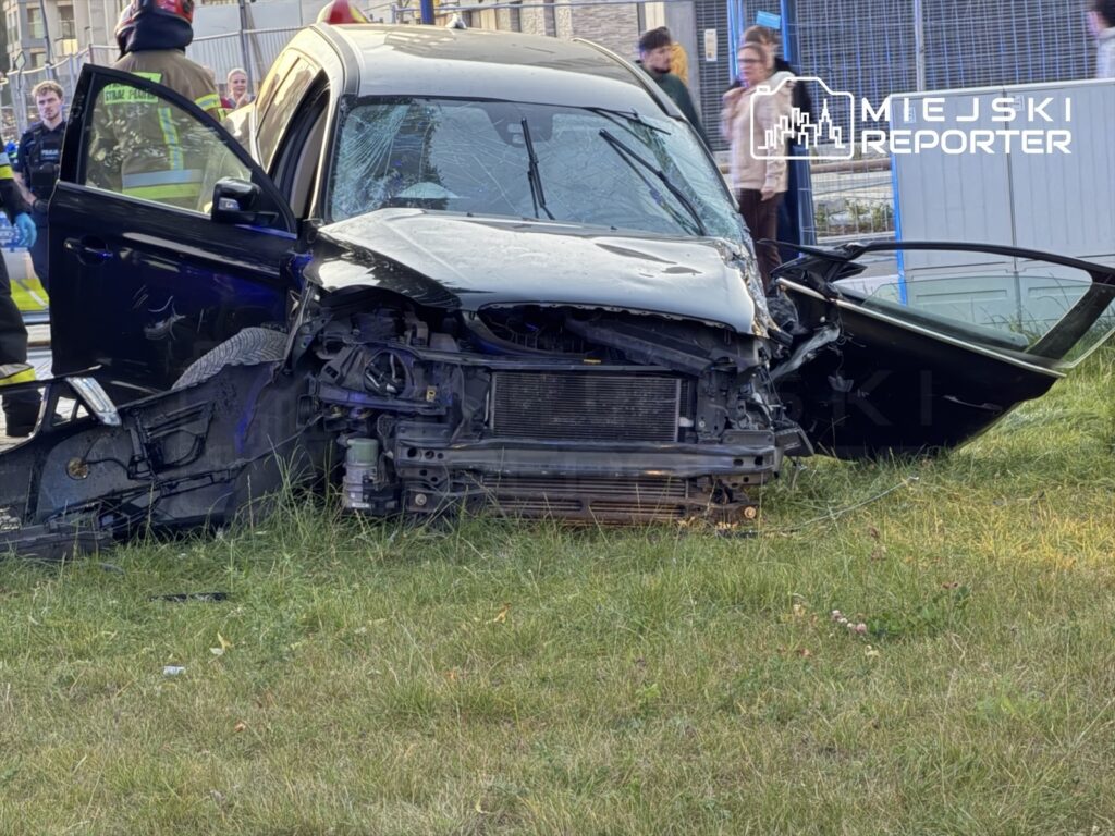 Rozbity samochód z otwartymi drzwiami stoi na trawniku, a uszkodzony przód i maska są widoczne na tle gapiów.