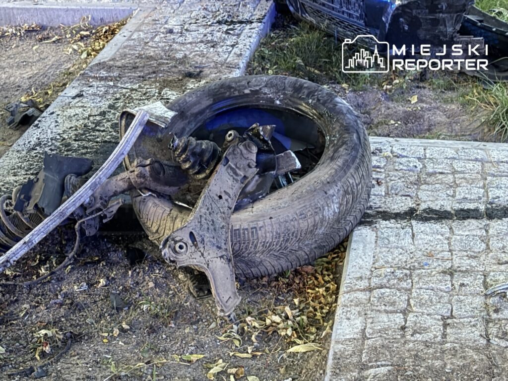 Uszkodzone koło i elementy zawieszenia samochodu leżą przy krawężniku na brukowanym chodniku.