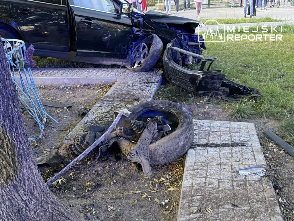 Uszkodzony czarny samochód z oderwanym kołem i zderzakiem stoi przy chodniku, obok leżą części auta.