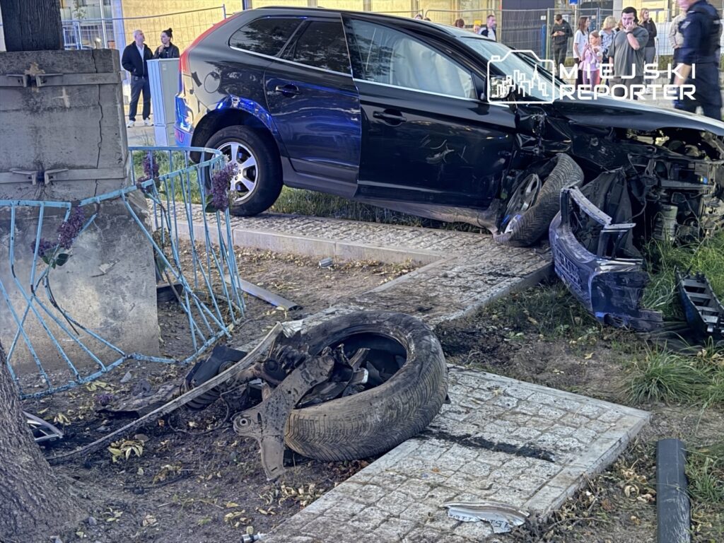 Ciemne kombi z rozlegle uszkodzonym przodem stoi przy murze; na ziemi leżą oderwane koło i części samochodu.