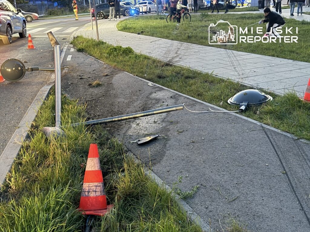 Przewrócony słupek i uszkodzona latarnia leżą na chodniku przy ulicy, obok których ustawiono pachołki drogowe.