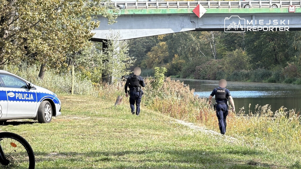 Dwóch policjantów idzie wzdłuż zarośniętego brzegu kanału, obok radiowozu i pod mostem.