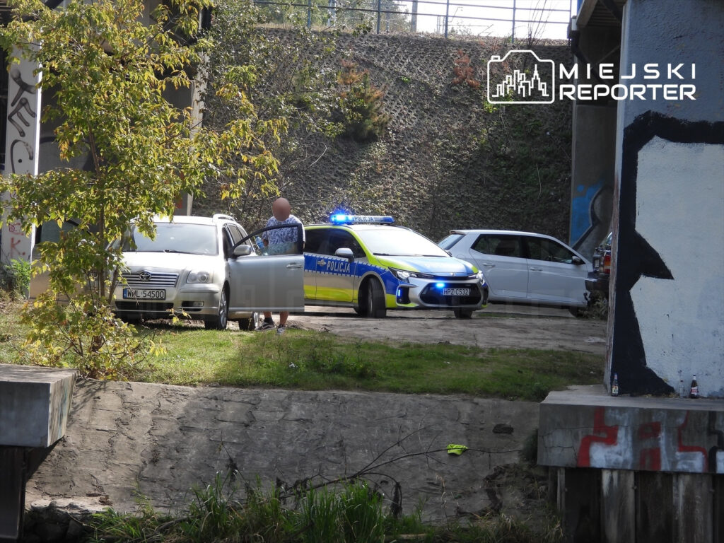 Radiowóz z włączonymi światłami stoi obok zaparkowanych samochodów pod wiaduktem, a policjant rozmawia z mężczyzną.