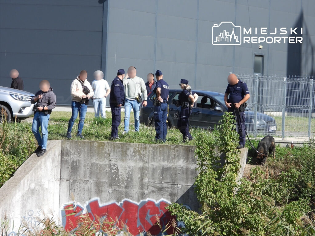 Policjanci i osoby cywilne stoją przy betonowym kanale, a policyjny pies węszy w zaroślach obok ogrodzenia.