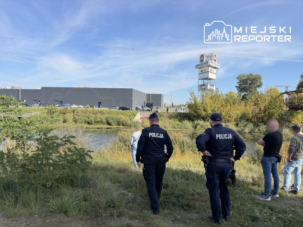 Policjanci i mężczyźni stoją nad porośniętym trzcinami kanałem, na tle przemysłowego budynku.