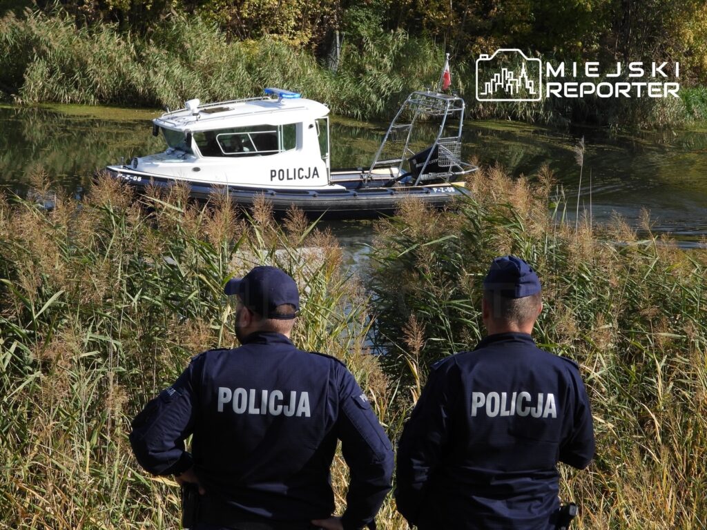 Dwóch policjantów obserwuje łódź policyjną na kanale, stojąc wśród trzcin.