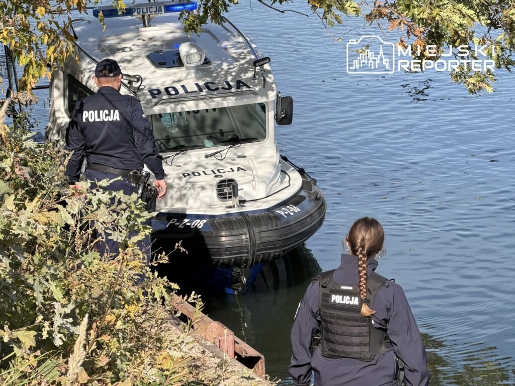 Policjanci stoją przy policyjnej łodzi cumującej przy zarośniętym brzegu kanału.