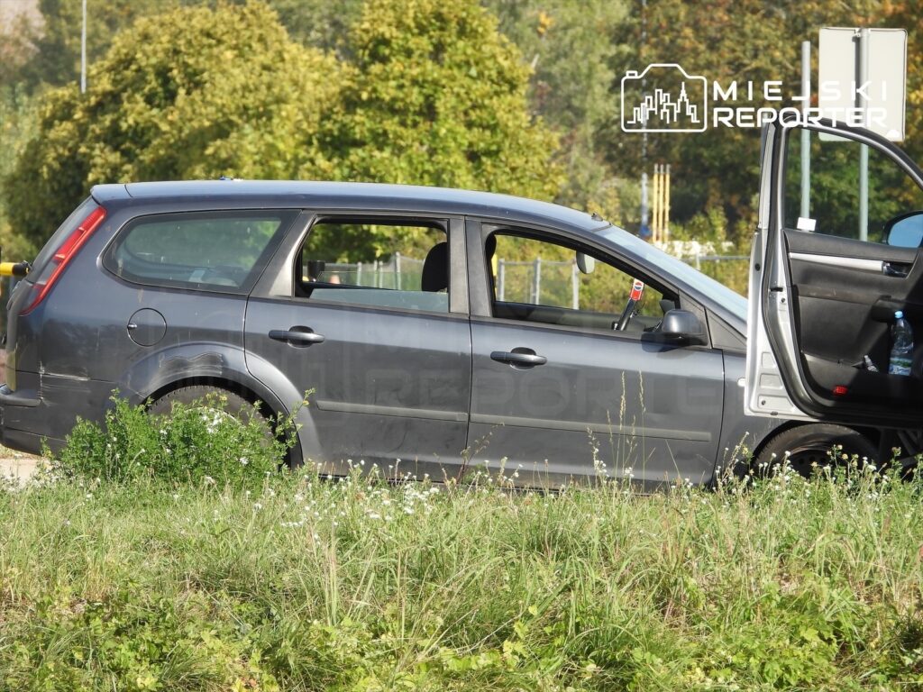 Szary samochód osobowy z otwartymi drzwiami stoi na zarośniętym poboczu przy ogrodzeniu.
