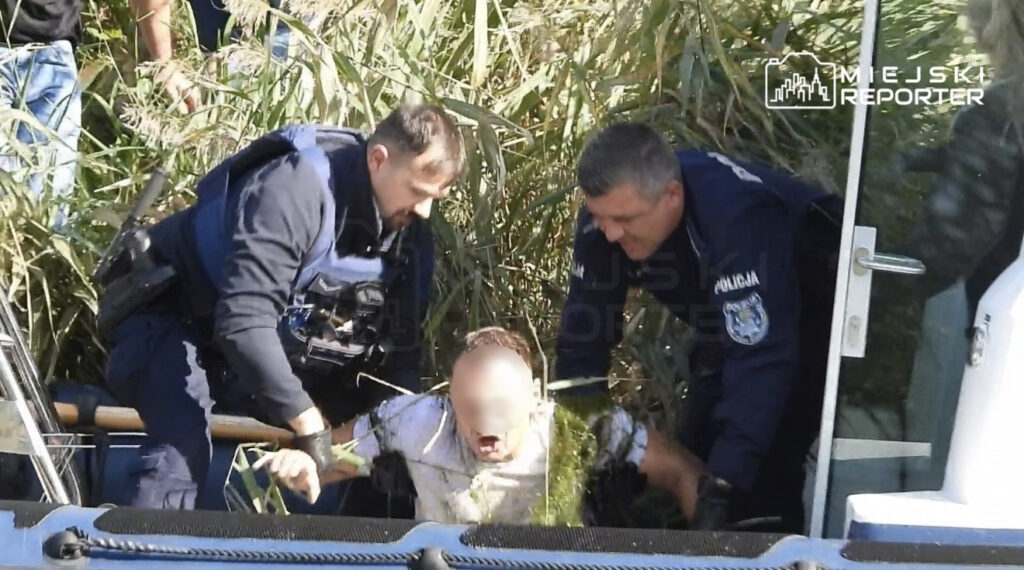 Dwóch policjantów wyciąga z trzcin mokrego mężczyznę na pokład łodzi przy kanale.