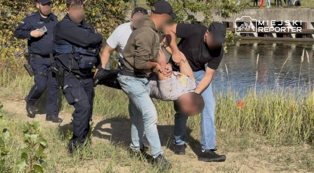 Policjanci i mężczyźni niosą zatrzymanego nad brzegiem kanału, wśród trzcin.