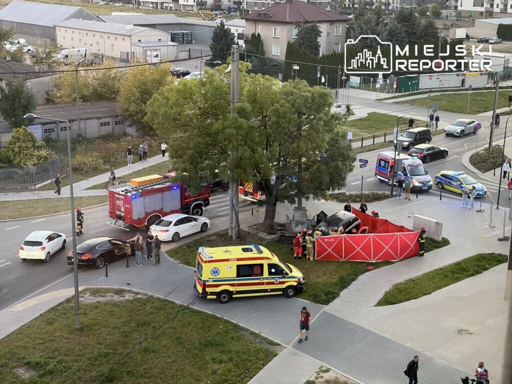 Strażacy i ratownicy medyczni przy wypadku na skrzyżowaniu, obok rozbitego auta i czerwonego parawanu.