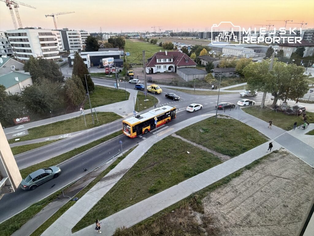 Samochody i autobus jadą przez skrzyżowanie wśród bloków mieszkalnych i placu budowy, widok z góry.