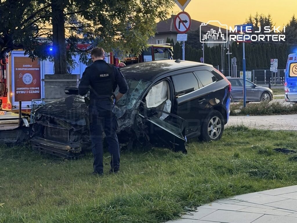Policjant ogląda mocno uszkodzone czarne kombi stojące na trawniku przy drodze, obok pojazdu służb ratunkowych.