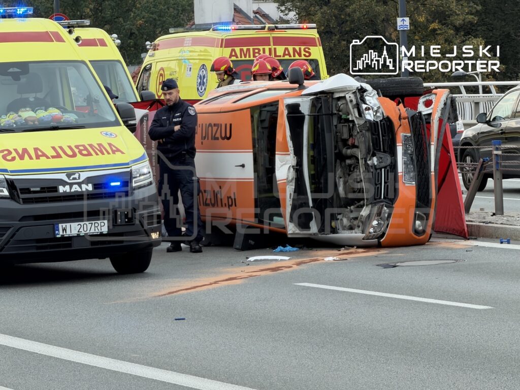 Przewrócona na bok pomarańczowa karetka na ulicy, obok stoją ratownicy medyczni i zaparkowane ambulanse.
