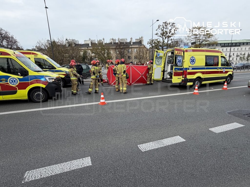 Strażacy w ubraniach ochronnych pracują przy czerwonym namiocie między karetkami na ulicy zabezpieczonej pachołkami.