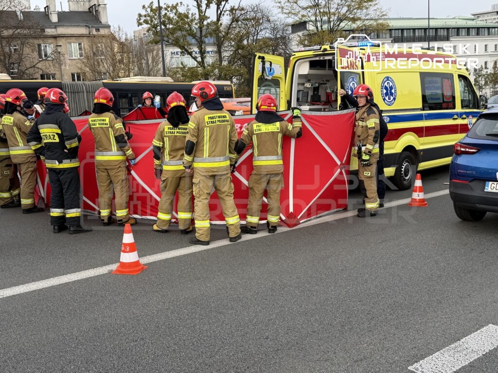 Strażacy w hełmach rozstawiają czerwoną osłonę przy karetce pogotowia na ulicy zabezpieczonej pachołkami.
