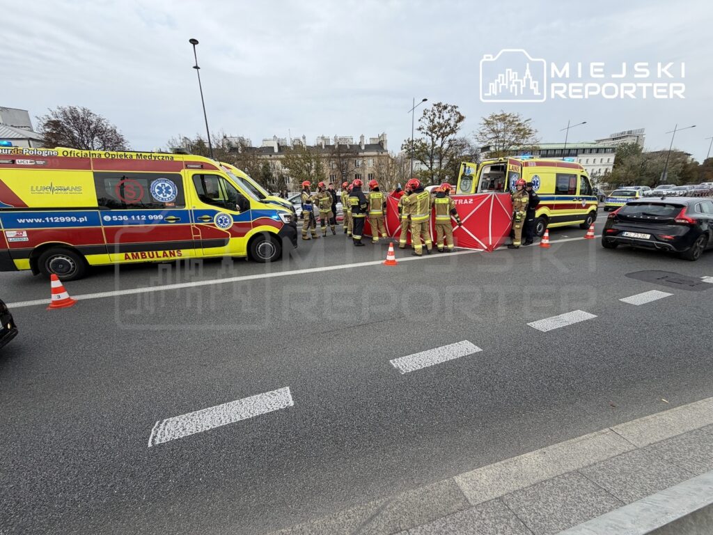 Ratownicy medyczni i strażacy zabezpieczają miejsce zdarzenia drogowego między ambulansami, za czerwonym parawanem.