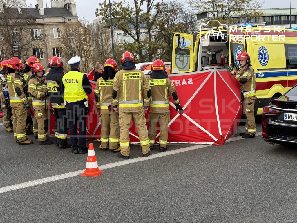 Strażacy i policjant osłaniają czerwonym parawanem miejsce zdarzenia przy karetce pogotowia na ulicy.