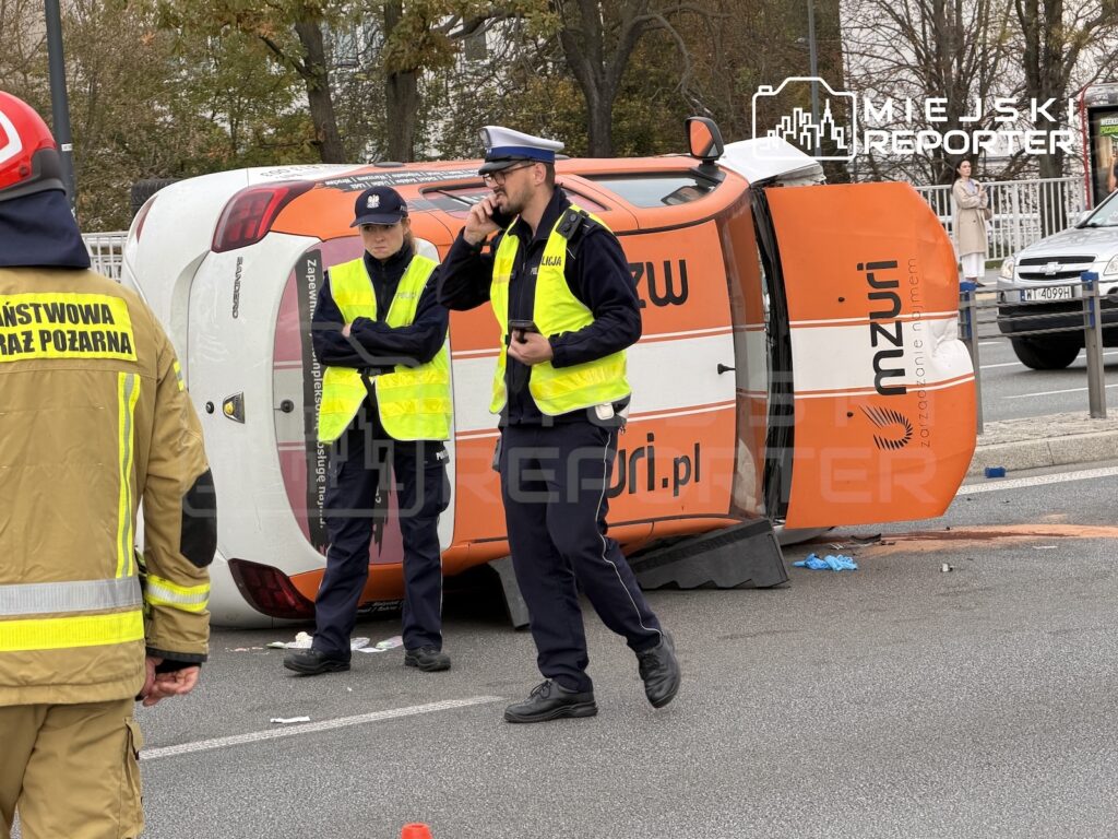 Policjanci i strażak zabezpieczają przewróconą pomarańczowo-białą karetkę na ulicy.