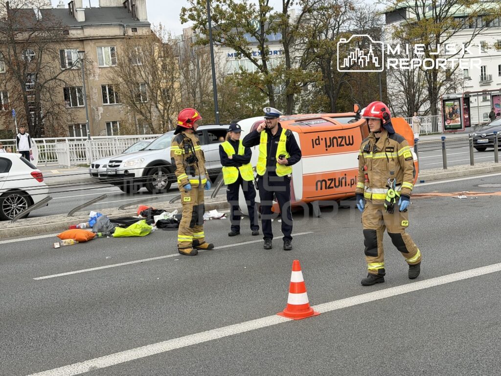 Strażacy i policjanci zabezpieczają miejsce wypadku na ulicy, obok leżącego poszkodowanego i samochodu dostawczego.