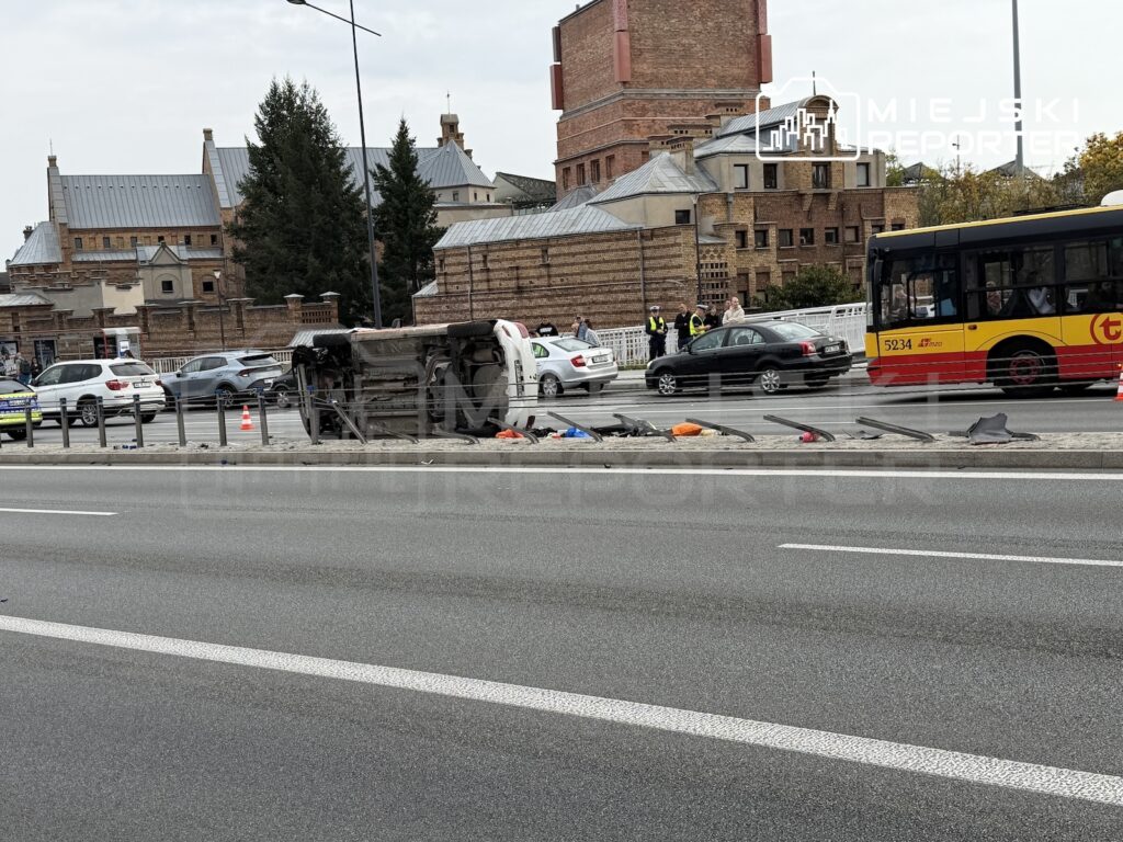 Przewrócony samochód leży na boku przy torowisku, obok stoją autobus, samochody i służby ratunkowe.