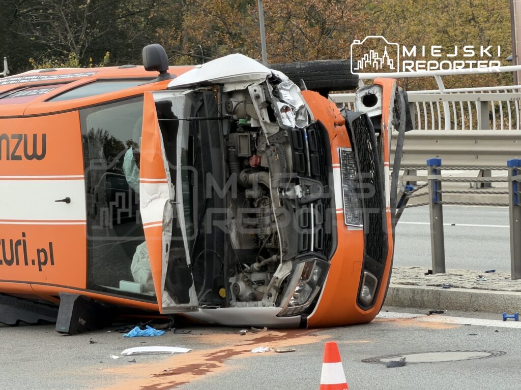Pomarańczowy samochód leży na boku na jezdni, z rozbitą przednią częścią i wyciekiem płynu na asfalt.
