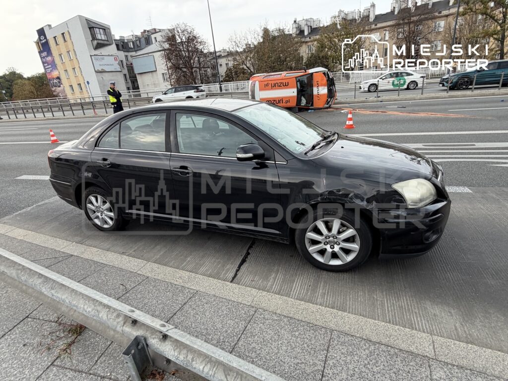 Czarny sedan stoi na jezdni przy przewróconej furgonetce, a drogę zabezpieczają pachołki.
