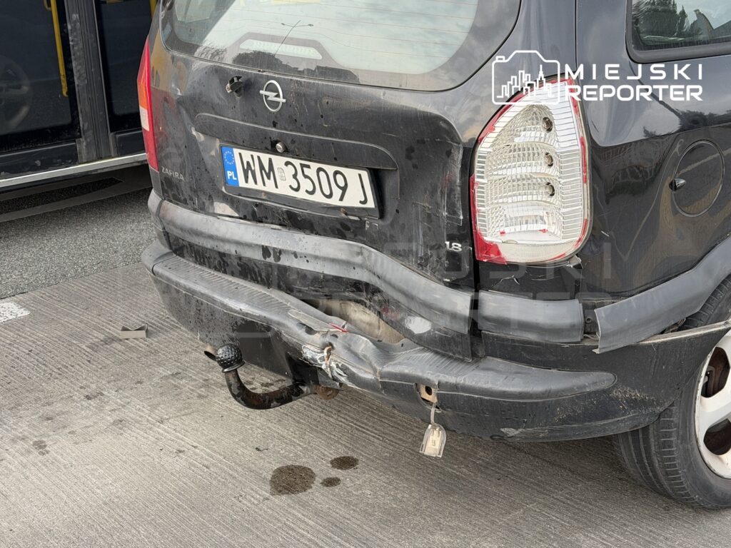 Tył szarego Opla Zafiry z uszkodzonym zderzakiem i hakiem holowniczym, zaparkowanego obok autobusu.
