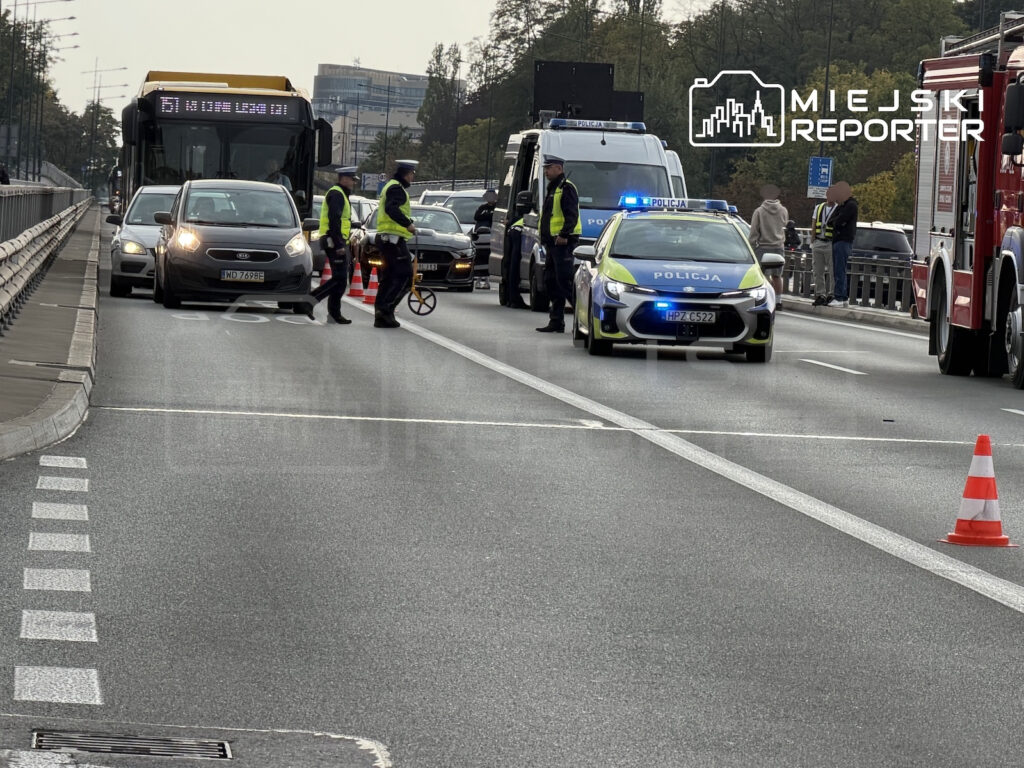 Policjanci w odblaskowych kamizelkach kierują ruchem przy radiowozie, autobusie i samochodach na zamkniętym pasie drogi.