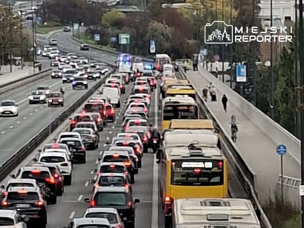 Korek samochodów i żółtych autobusów na wielopasmowej drodze, obok której biegnie chodnik z pieszymi.