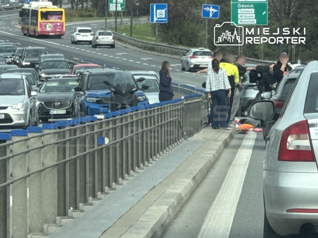 Piesi stoją przy leżącej osobie na pasie drogi między zakorkowanymi samochodami.