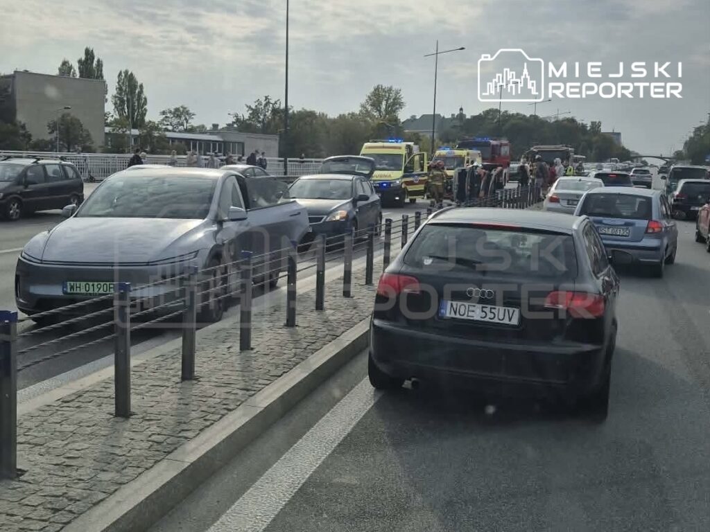Samochody stoją w korku na ulicy, a za nimi widać pojazdy służb ratunkowych i grupę ludzi.