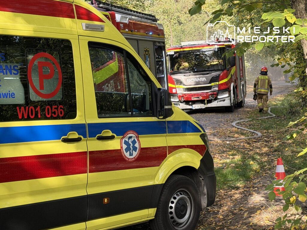 Karetka pogotowia i wóz strażacki stoją na leśnej drodze, a strażak idzie wzdłuż rozwiniętego węża gaśniczego.