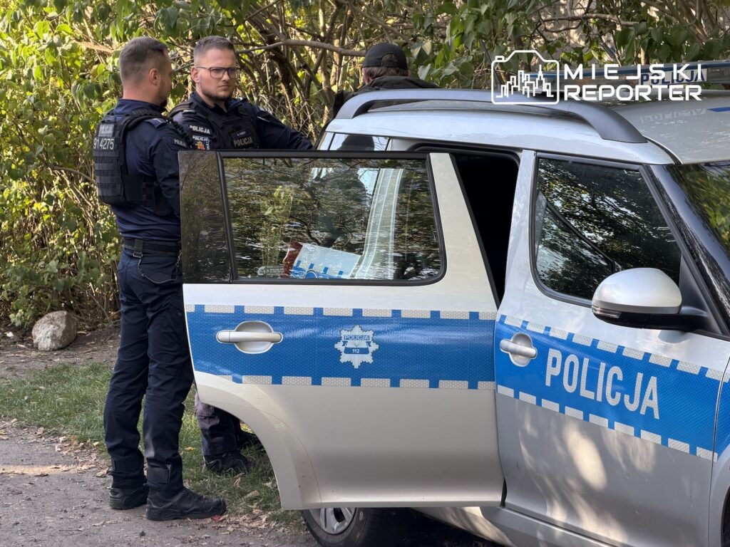 Dwóch policjantów stoi przy otwartych drzwiach radiowozu na zalesionej drodze.