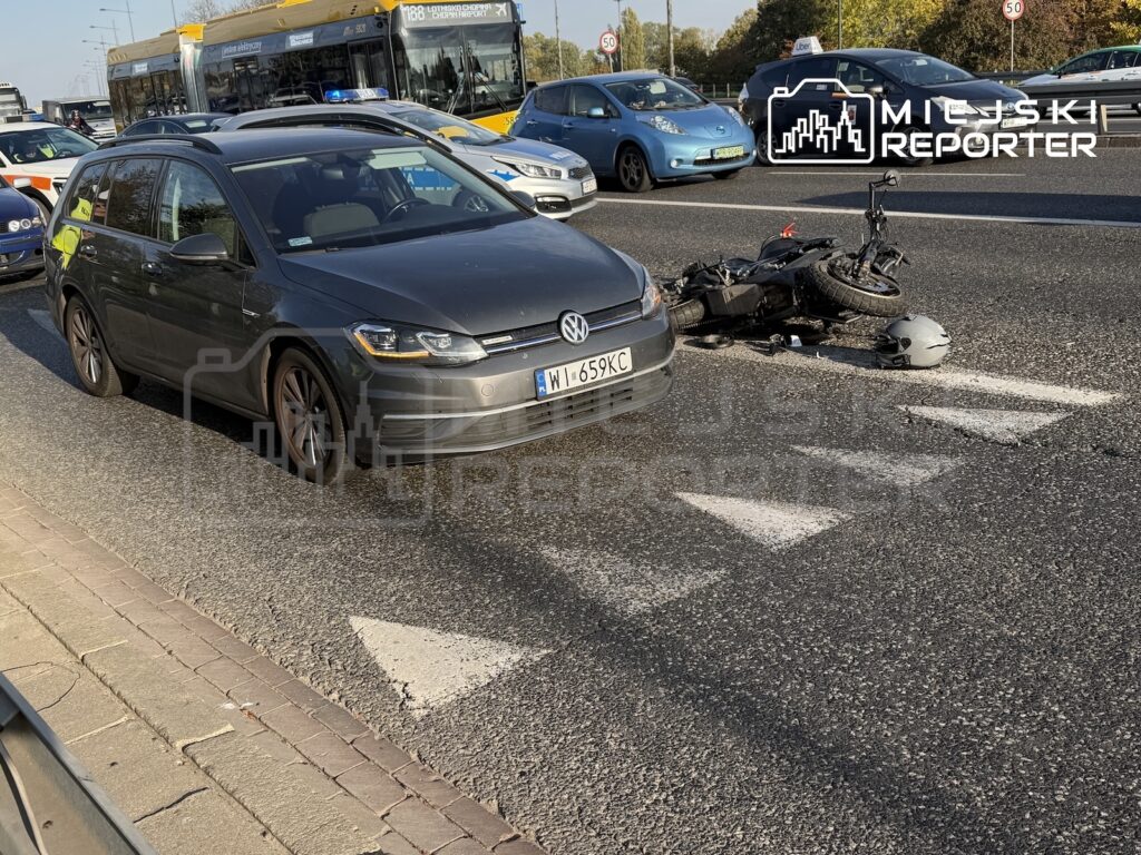 Przewrócony motocykl i kask leżą na przejściu dla pieszych obok ciemnego samochodu, wśród miejskiego ruchu ulicznego.