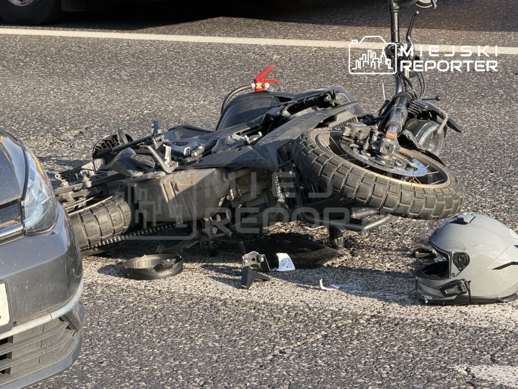 Uszkodzony motocykl leży na jezdni między samochodem a kaskiem, obok widać rozrzucone części.