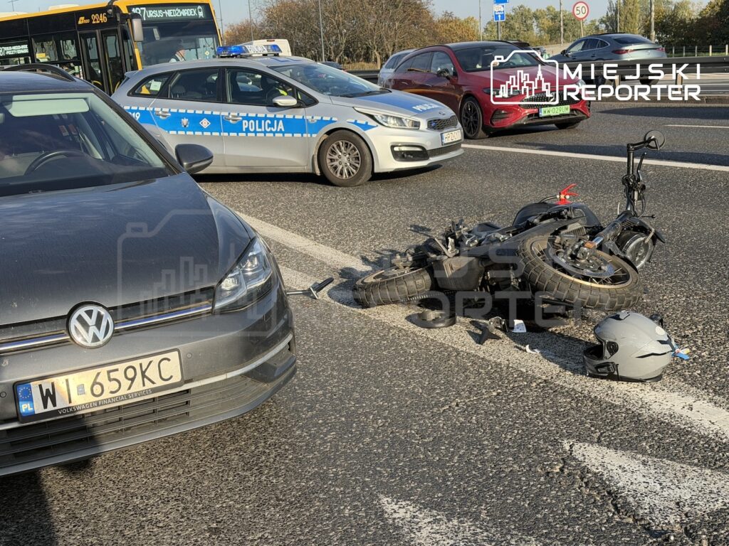 Przewrócony motocykl i kask leżą na jezdni obok samochodu, a policja blokuje pas ruchu.