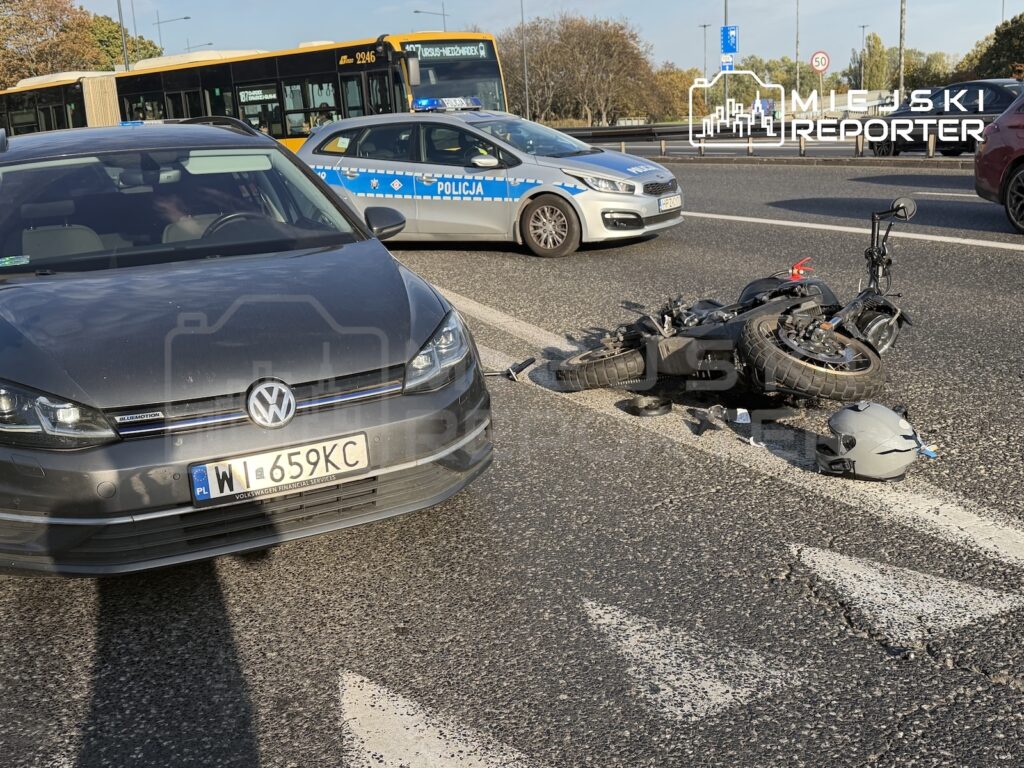 Przewrócony motocykl i kask leżą na jezdni obok samochodu, a w tle stoi policyjny radiowóz i autobus.