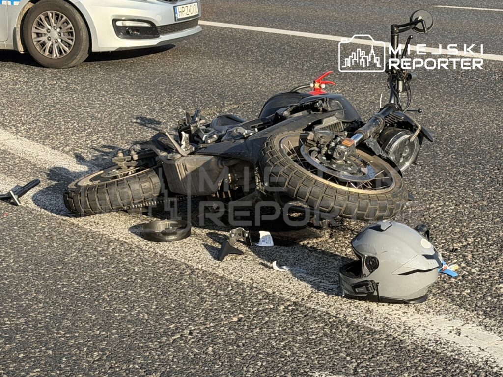 Przewrócony motocykl i leżący obok kask na jezdni, obok widoczny przód białego samochodu.
