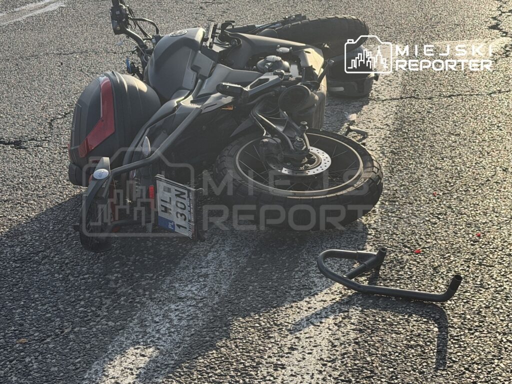 Motocykl leży przewrócony na asfaltowej jezdni, obok leży oderwany element pojazdu.