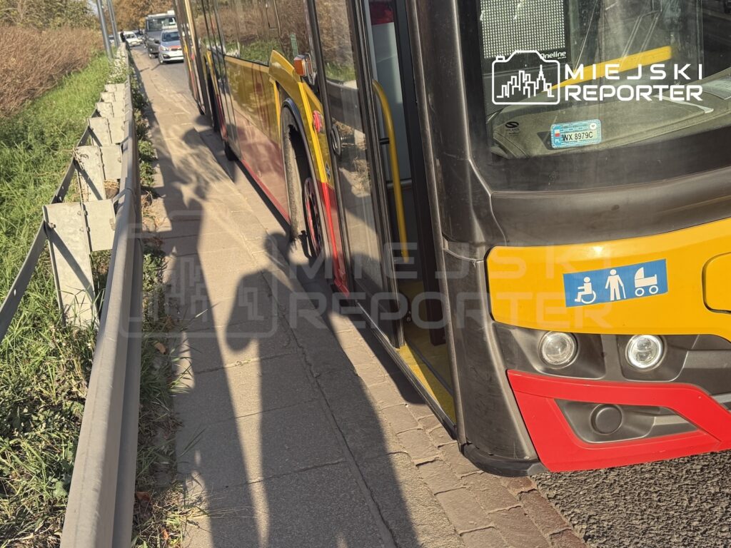 Tył żółto-czerwonego autobusu miejskiego stojącego przy wąskim chodniku oddzielonym metalową barierką od jezdni.
