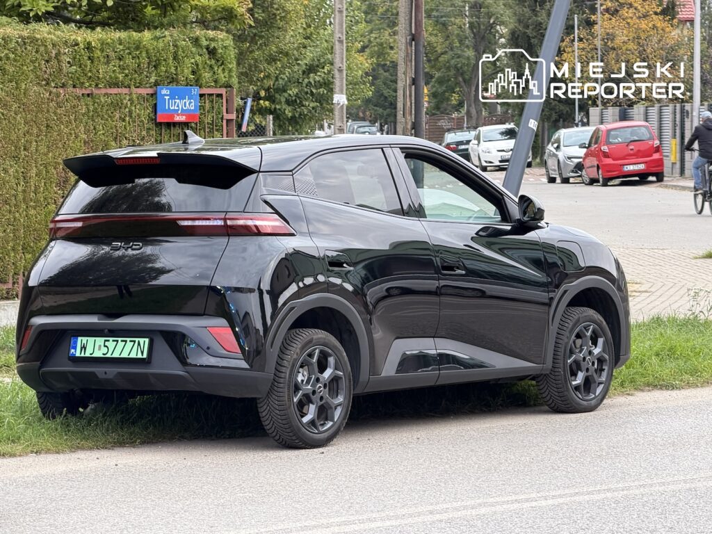 Czarny samochód elektryczny BYD zaparkowany przy ulicy, obok znaku przystanku „Tużycka”.
