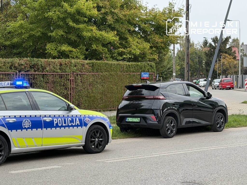 Czarny samochód elektryczny zaparkowany przy radiowozie na ulicy, obok chodnika i ogrodzenia.