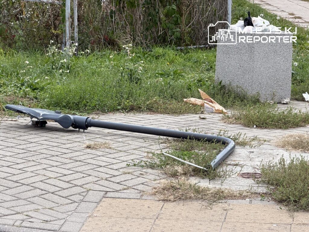 Przewrócona latarnia leży na chodniku z kostki brukowej, obok zarośniętego trawnika.