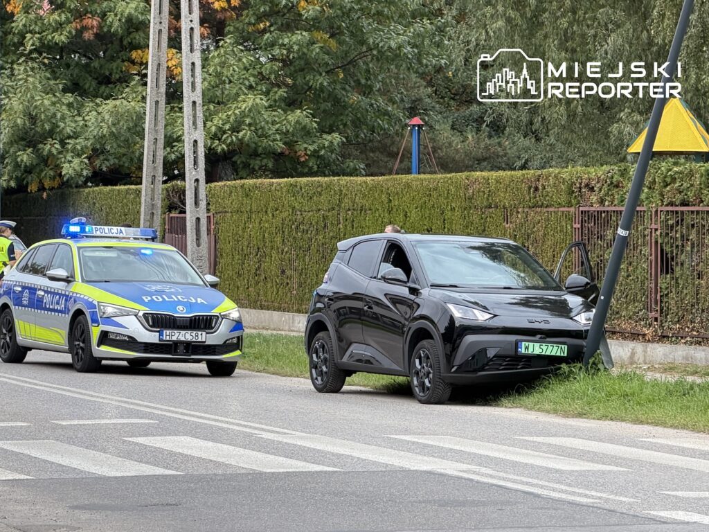 Czarny samochód BYD stoi przy skrzyżowaniu, obok radiowozu policji i pochylonej latarni.