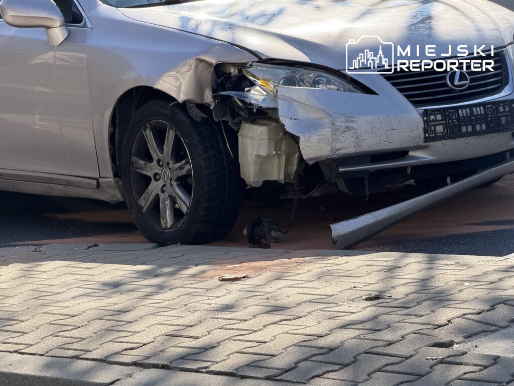 Srebrny Lexus z uszkodzonym przednim zderzakiem i reflektorem stoi przy chodniku, na kostce leżą odłamki.