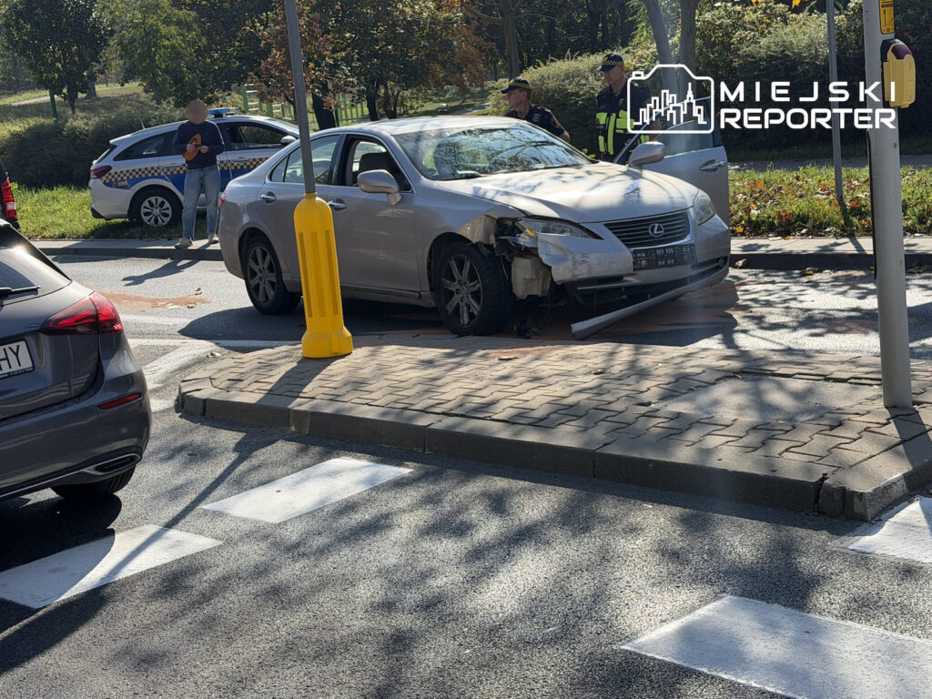 Srebrny Lexus z uszkodzonym przodem stoi na chodniku przy przejściu dla pieszych, obok policjanci i radiowóz.