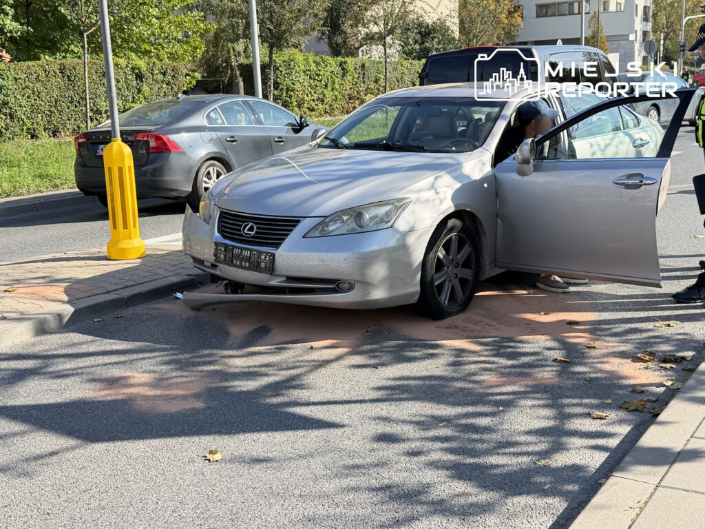 Srebrny Lexus stoi na jezdni z otwartymi drzwiami, obok zaparkowanego samochodu i żółtego słupka.